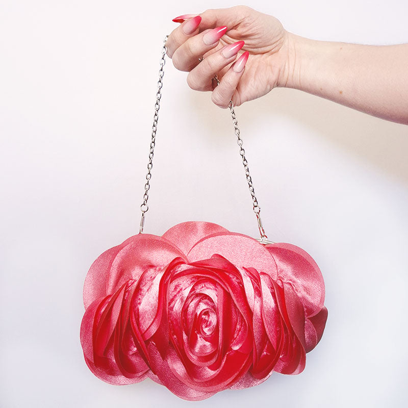 An elegant close-up of hands holding the Rose Chic Petal Clutch by its delicate chain strap. A delicate rose-shaped clutch bag with layered petal detailing in a soft, romantic hue. The elegant floral design mimics the texture of blooming petals, creating a timeless statement piece. Set against a refined backdrop, the clutch exudes femininity and sophistication, perfect for weddings, cocktail events, and special occasions. Rose Chic Petal Clutch Bag in rose pink by Montipi Australia
