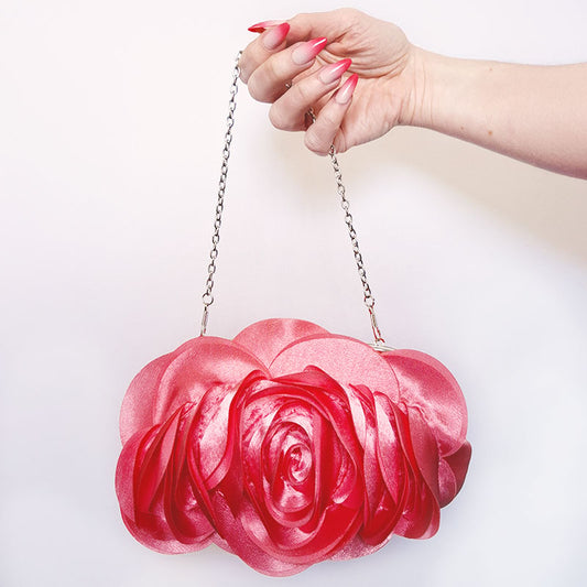 An elegant close-up of hands holding the Rose Chic Petal Clutch by its delicate chain strap. A delicate rose-shaped clutch bag with layered petal detailing in a soft, romantic hue. The elegant floral design mimics the texture of blooming petals, creating a timeless statement piece. Set against a refined backdrop, the clutch exudes femininity and sophistication, perfect for weddings, cocktail events, and special occasions. Rose Chic Petal Clutch Bag in rose pink by Montipi Australia
